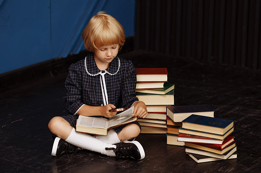 A schoolgirl sits on the floor with an open book surrounded by textbooks. The concept of studying, homework. For projects about education, school competitions.