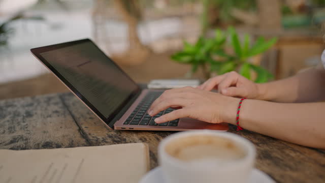 Hands of young woman coding on laptop in outdoor seaside cafe. Female freelancer typing on computer at tropical location by ocean. Young adult working remotely on exotic island. Worldwide work concept