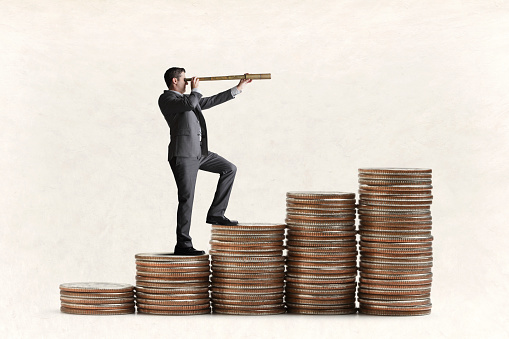 A businessman stands on a row of coin stacks as he peers through a spyglass toward his financial future.