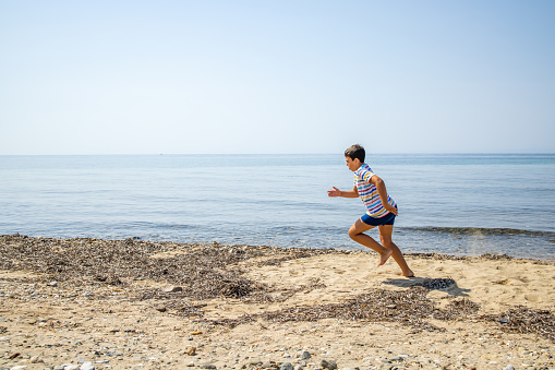 Side-view of Caucasian boy in shirt and swimwear running on the beach during sunny morning