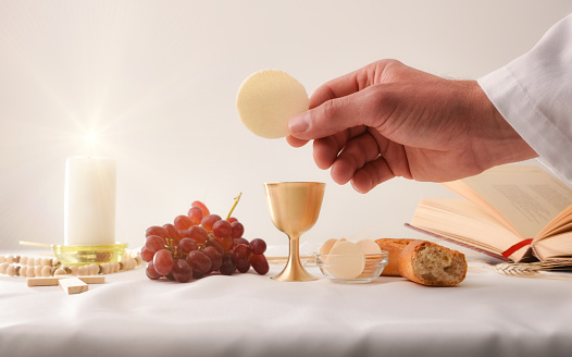 Hands of a priest consecrating a host as the body of Christ to distribute it to the communicants with table with sacred objects behind.