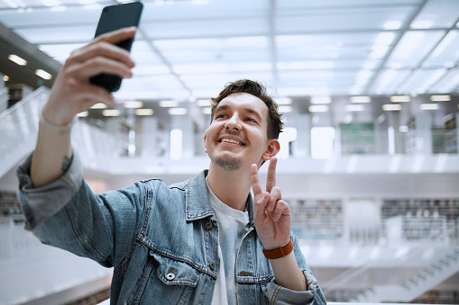 Selfie, university and man student with peace sign for social media update, blog post or live streaming his experience. Education, research and person in campus library with smile for profile picture