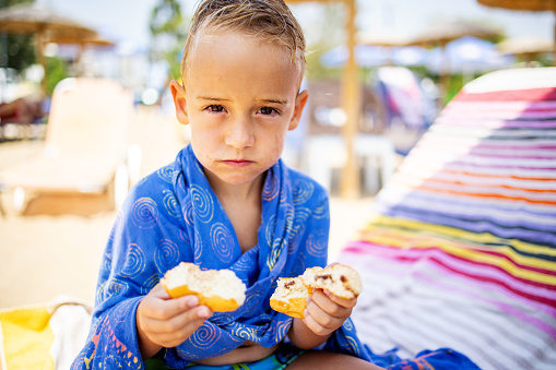 Portrait of a young Caucasian boy, wrapped in a towel, sitting on the beach chair, and eating pastry