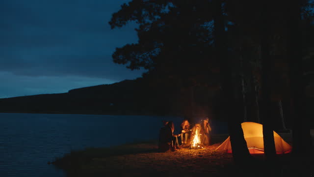 group of friends sitting by campfire roasting marshmallows camping in forest by lake an night chatting sharing warmth enjoying outdoor adventure 4k