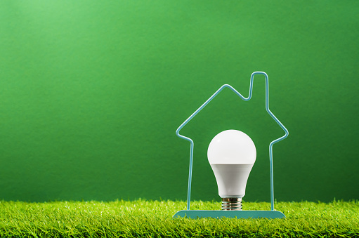 Glowing LED light bulb inside house frame on green grass background. Green energy saving and smart house solutions. Sustainable consumption. Earth Day