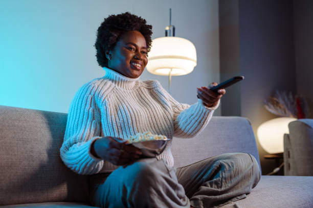 mulher afro-americana assistindo programa de tv favorito e comendo pipoca em casa - entertainment home interior electronics industry remote control - fotografias e filmes do acervo