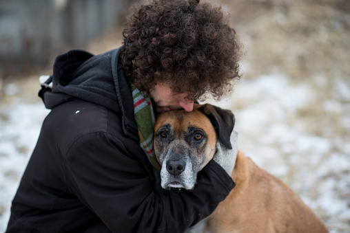 Young Caucasian man wearing a winter coat and a scarf hugging his emotional support dog lovingly to comfort him as they are out for a walk through a snowy forest during winter. The dog is a mixed breed and brown and black in color with a sad expression on his face.