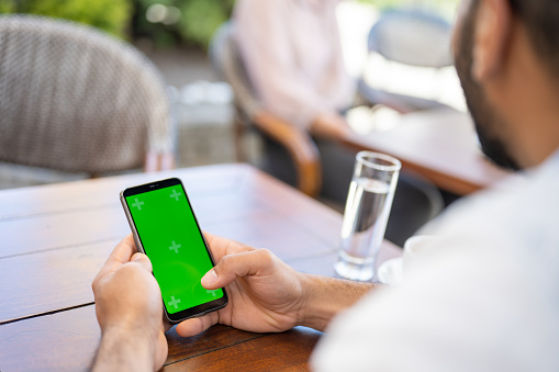 Man sitting at outdoors cafe table and using smartphone with green screen close up