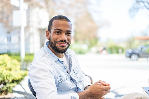 Smiling multiracial man looking at camera while sitting on bench on city street at day time