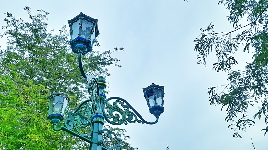 This stunning image showcases a city park's lamp during daylight hours, captured from a low angle with a green foliage background and a clear blue sky. The lamp's intricate design and warm glow provide an inviting atmosphere that complements the natural beauty of the park. This photo is perfect for advertising campaigns, website banners, or any project that requires a visually appealing and engaging cityscape.