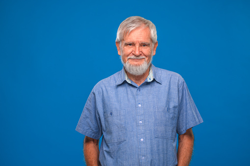 Senior man portrait with small smile in blue background. Adult with grey hair, beard and mustache looking at camera.