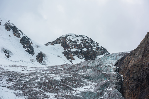 Dramatic landscape with large icefall on glacier under snow mountain peak in gray cloudy sky. Sharp rocks and beautiful glacier with icefall in high altitude. Gloomy scenery in mountains in overcast.