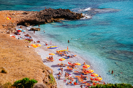 The western coast of Sicily, the Mediterranean Sea. A pebble beach full of people and lined with colorful umbrellas.