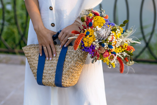 A girl in a white dress holds wild flowers in a wicker straw bag. Vintage eco style concept. Stylish wicker straw bag with dried wildflowers close-up