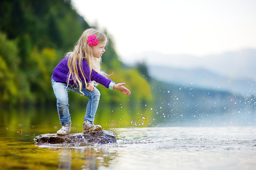 Adorable girl playing by Hallstatter See lake in Austria on warm summer day. Cute child having fun splashing water and throwing stones into the lake. Summer activities for kids.