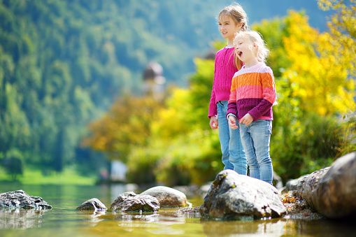 Adorable sisters playing by Konigssee lake in Germany on warm summer day. Cute children having fun feeding ducks and throwing stones into the lake. Summer activities for kids.