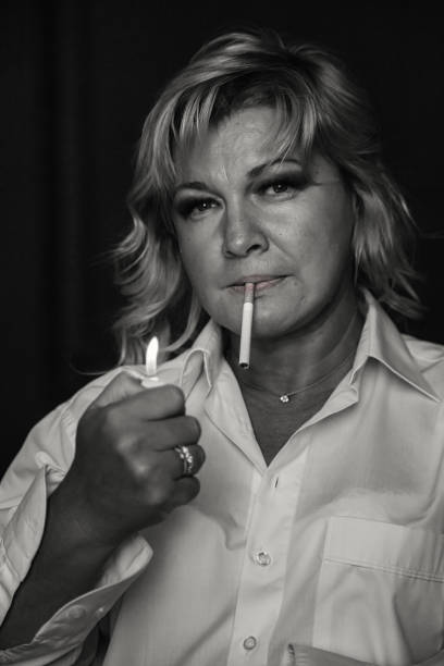 black white portrait of middle aged 45 years old woman lighting cigarette at black background. thoughtful sad alone mature lady in white, aging image. loneliness emotion concept. copy ad text space - women middle aging process 45 50 years imagens e fotografias de stock