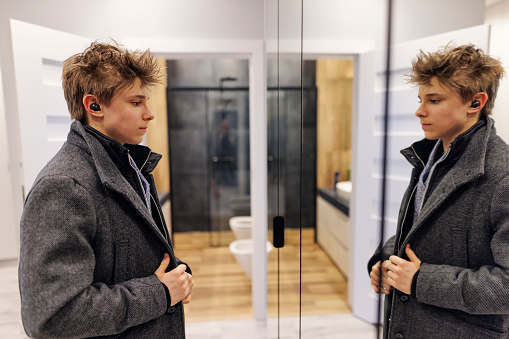 Teenage boy is preparing to go out. The boy is listening to the music using wireless in-ear headphones. The boy is putting on a coat and looking at the mirror.\nShot with Canon R5