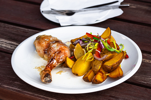 Chicken leg with baked potato with vegetable circle and cream sauce on white plate, cutlery on wooden table.