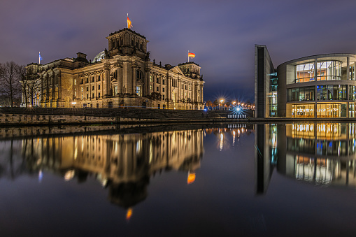 View of government buildings at night time. Reichstag in Berlin at the blue hour. Illuminated historical buildings. River Spree with reflection on water surface in winter