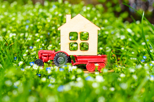 Miniature tractor carrying a flat house model placed on a flowerbed of lush green grass and colorful spring flowers. Springtime as a season for a new beginning.