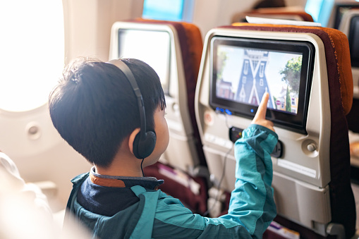 In-flight technology creates enjoyment during the journey. Back view of Asia boy wearing headphone and jacket sitting on the plane touching screen of digital tablet to choose cartoon during flight.