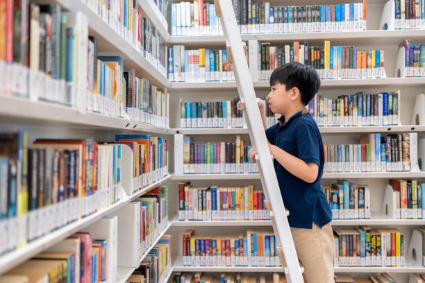 czytanie książek to nieskończony wzrost wiedzy. asia chłopiec wybiera podręcznik w bibliotece w szkole podstawowej. chłopiec wspiął się po drabinie w bibliotece, aby znaleźć książkę. baw się dobrze i ciesz się czytaniem książki. koncepcja - male education college student nerd zdjęcia i obrazy z banku zdjęć