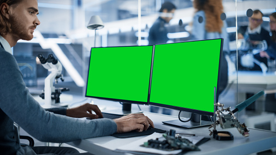 Software Developer Working on Computer with Green Screen Mock Up Display. Scientific Lab, Engineering Research Center with Specialists Working on Mobile Robot Development in the Background.