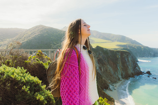 Portrait of happy female with long hair wearing colorful knitted sweater exploring the scenic mountain coast with old bridge on the background on Monterey peninsula in the USA during sunny spring day