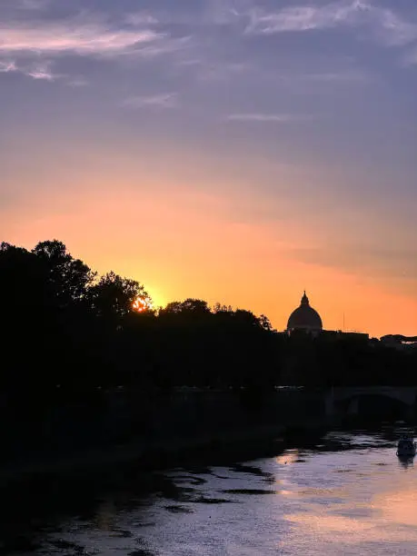 The photo shows the sunset over the Tiber in Rome, with the silhouette of Cupolone di San Pietro in the distance. The sky fades from red to orange.