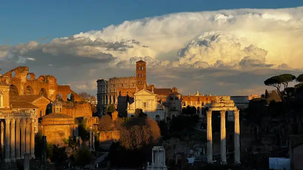Image of Ancient Rome at sunset, with a huge cloud in the sky. The remains of the Colosseum and the Roman Forum create a striking contrast with the cloud.