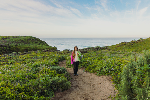 Front view of female with long hair wearing colorful knitted sweater walking at the scenic footpath on the flowering meadow to the ocean on Monterey peninsula in the USA during sunny spring day