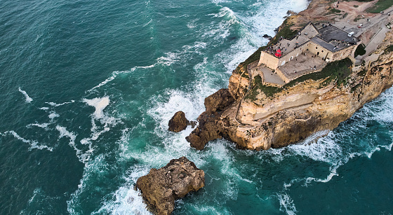 Aerial view of an old lighthouse on a cliff with a fortress on the coast of the Atlantic ocean in Nazare town, Portugal. High quality photo