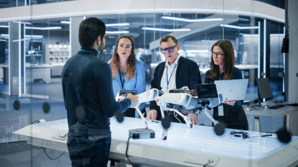 istock Diverse Team of Industrial Robotics Specialists Gathered Around a Table With a Mobile Robot. Scientists Use Tablet and Laptop Computers to Program the Automated AI Robotic Dog. 1482153314
