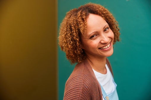 Portrait of a woman with curly hair standing sideways and smiling outside in front of a colorful wall
