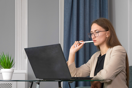 Young, beautiful, confident woman in glasses and a light jacket is sitting at a table and attentively looking at a laptop, holding a pen near her lips. Concept of remote work, online learning