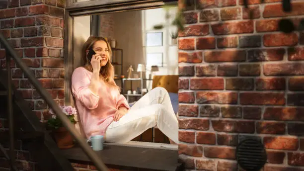 Beautiful Young Female Having a Call on her Smartphone While She Sits on her Windowsill. Expressive Woman Speaking on the Phone, Talking to her Long-Distance Partner on Sunday Evening.
