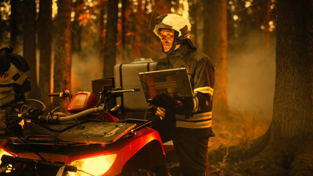 mutiger geschickter feuerwehrmann, der neben einem atv steht und einen laptop im wald mit wütendem buschfeuer benutzt. superintendent oder squad leader stellt sicher, dass die notfallsituation unter kontrolle ist. - fire department courage forest fire heroes stock-fotos und bilder