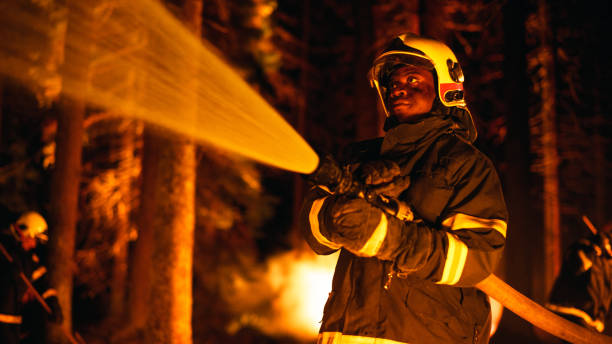 ruhiger und gesammelter afroamerikanischer feuerwehrmann löscht einen waldbrand tief in einem wald. profi in sicherheitsuniform und helm mit einem feuerwehrschlauch, um gefährliche waldbrände zu bekämpfen. - fire department courage forest fire heroes stock-fotos und bilder