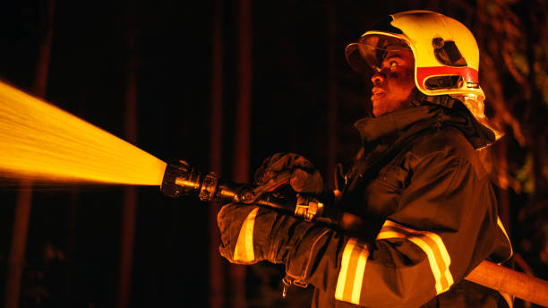 침착하고 집중된 아프리카계 미국인 소방관이 숲 속 깊은 곳에서 산불을 진압하고 있습니다. 위험한 산불과 싸우기 위해 소방 호스를 사용하는 안전 유니폼과 헬멧을 쓴 전문가. - fire department courage forest fire heroes 뉴스 사진 이미지