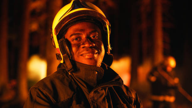 안전복을 입은 용감하고 잘생긴 젊은 성인 소방관의 초상화와 산불 동안 카메라를 향해 포즈를 취하는 헬멧을 클로즈업합니다. 전문 흑인 소방관이 카메라를 보고 웃고 있다. - fire department courage forest fire heroes 뉴스 사진 이미지