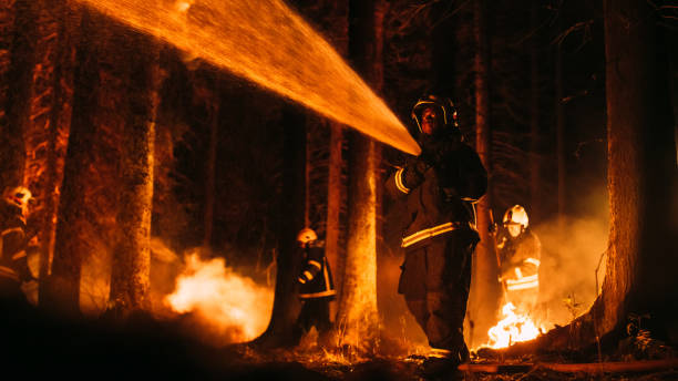 istock Experienced African American Firefighter Extinguishing a Wildland Fire in a Forest. Professional in Safety Uniform and Helmet Using a Fire Hose to Battle Dangerous Wildfire. Team is Working Behind. 1482174067