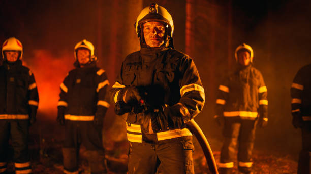 porträt eines tapferen, gutaussehenden besatzungsführers in sicherheitsuniform und helm, der während eines lauffeuers mit feuerwehrleuten für die kamera posiert. berufsfeuerwehrmann mit einem feuerwehrschlauch im arm. - fire department courage forest fire heroes stock-fotos und bilder