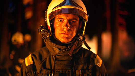 Portrait of a Brave Handsome Young Adult Firefighter in Safety Uniform and a Helmet with a Flashlight. Professional Fireman Looking at Camera. Wildland Fire in a Forest in the Background.