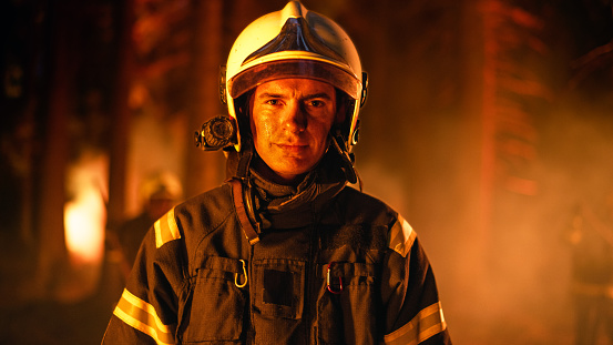 Retrato de un bombero adulto joven seguro y guapo con uniforme de seguridad y un casco con una linterna. Bombero profesional mirando a la cámara. Incendio forestal en un bosque al fondo. photo