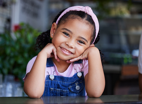 Black child, face and girl portrait with a smile, happiness and cute clothes with hands for headshot. Happy kid at a table for fashion, positive mindset and blurred background at cafe or home