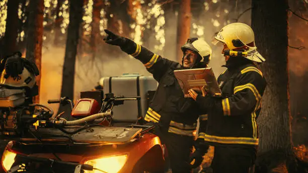 Photo of Portrait of Two Professional Firefighters Standing Next to ATV, Discussing the Situation During a Wildland Fire: Brave Superintendent Talking with Experienced Squad Leader, Using Laptop Computer.