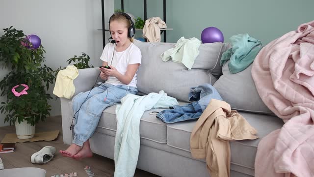 A girl is playing on a mobile phone. The room is a mess. The child scattered things in the room.