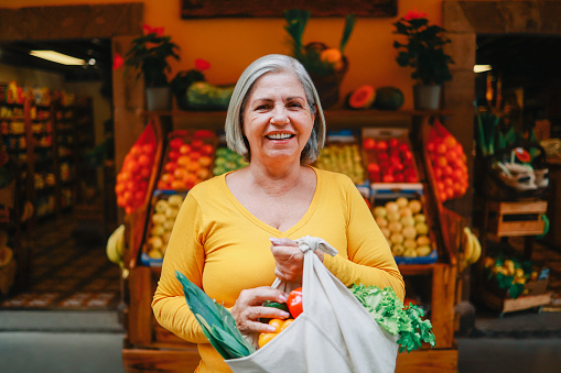 Zero waste food retail - Happy senior woman holding reusable bag of fresh vegetables at local market store - Sustainable eco concept - Focus on face