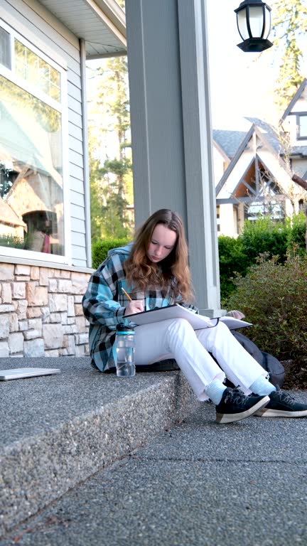 girl draws with pencil in large notebook notebooks camera slowly pulls back filming country house bottle of water white jeans black sneakers checkered blue jacket long brown hair of teenage girl